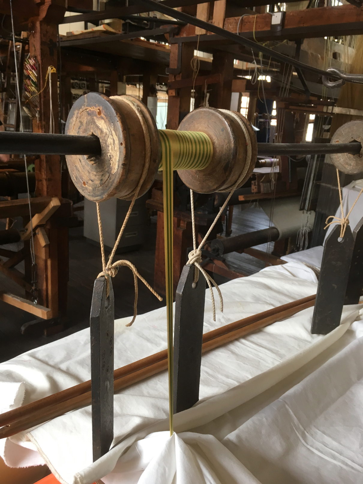 Photograph of a bobbin hanged up in an antique loom, weighted on both sides, with a ribbon's worth of silk wound on it.