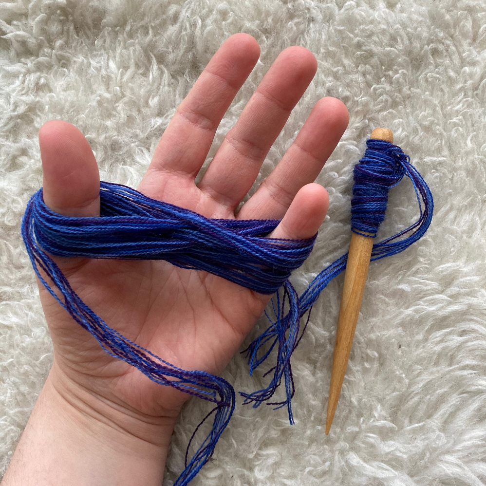 A hand holding a freshly wound yarn butterfly, next to a wooden tapestry bobbin filled with purple yarn.