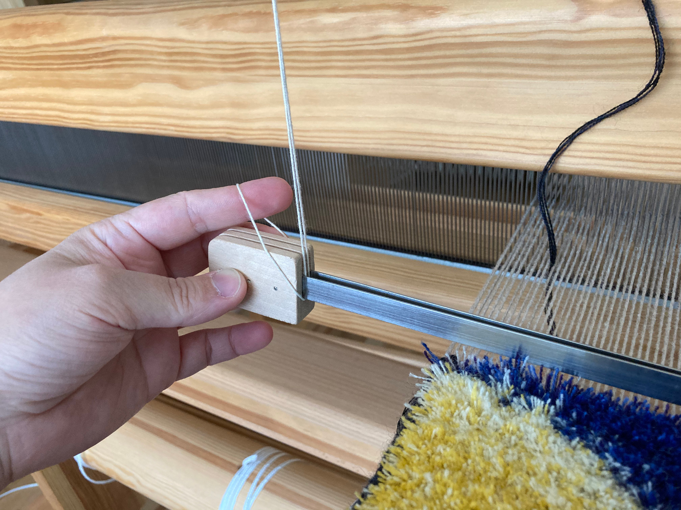 A hand lightly holding a flossa ruler suspended at the front of a loom, above an in-progress carpet weave.